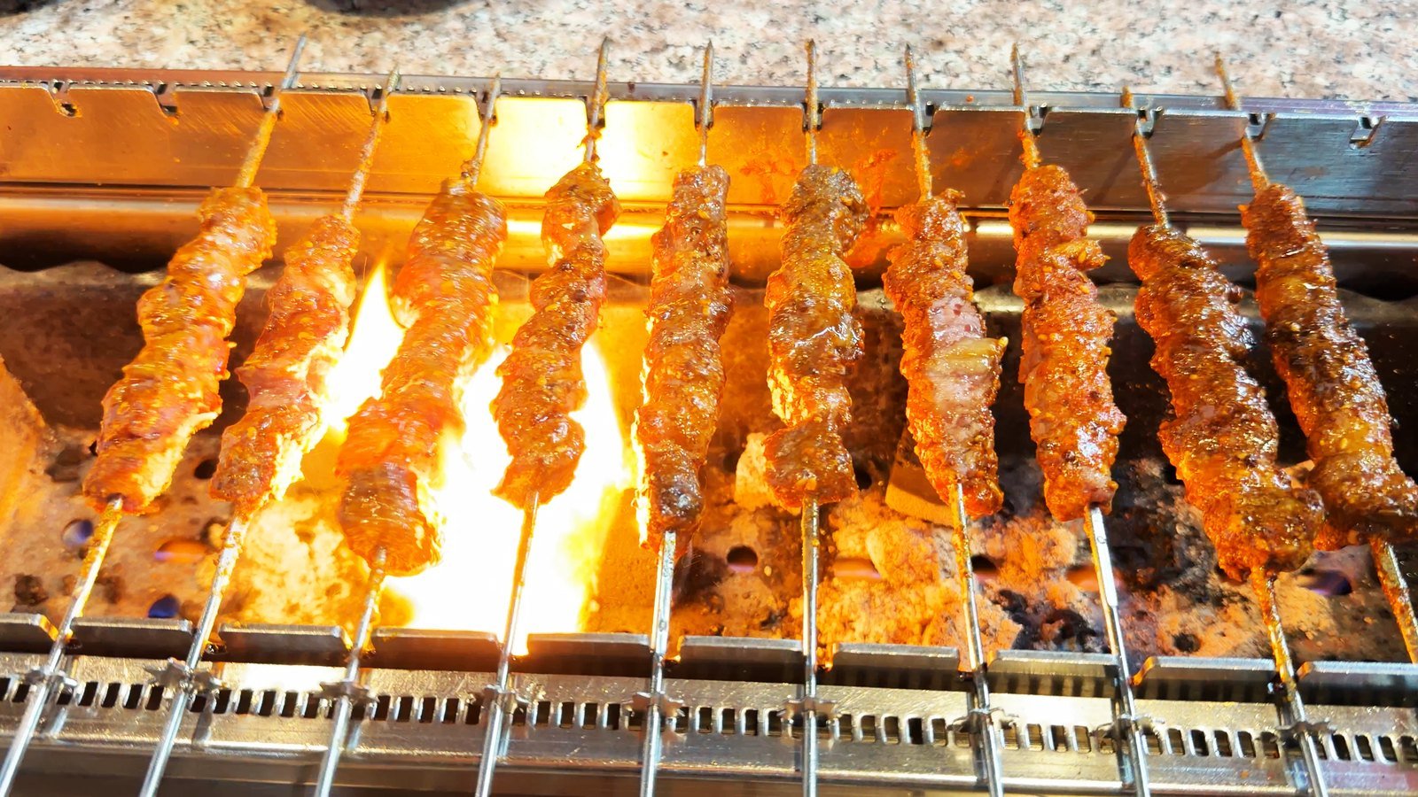 Rows of cumin-crusted lamb skewers browning over an open flame on a trough grill at a lamb skewer restaurant in Los Angeles Koreatown